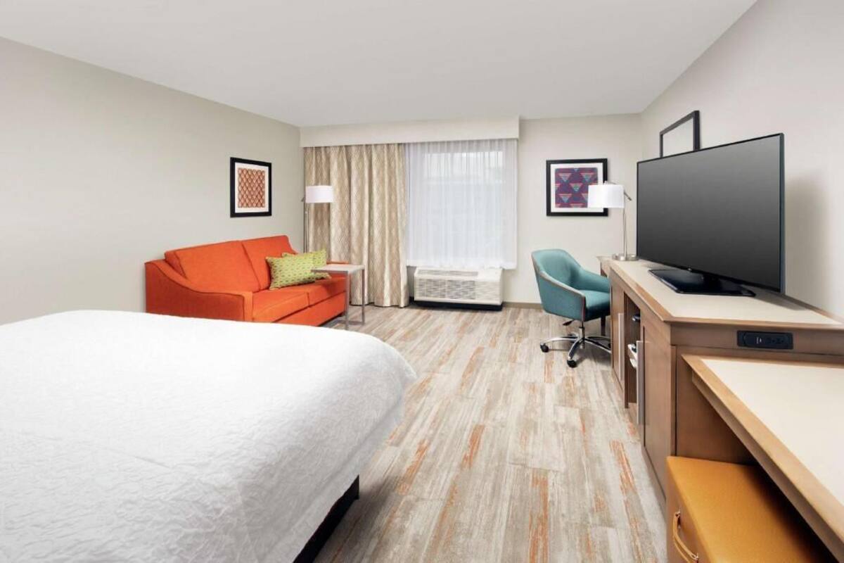 A hotel room with a bed, a desk with a flat-screen TV, a chair, a sofa, and a window with curtains.