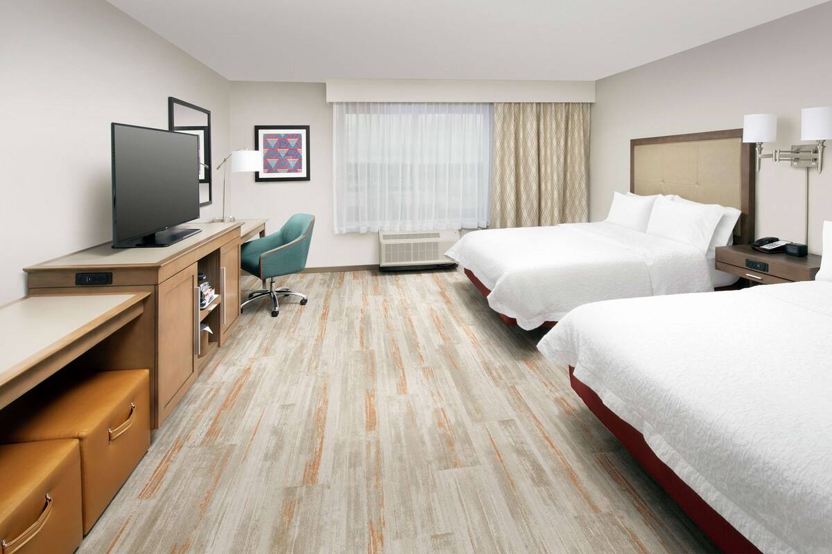 A hotel room with a desk, two beds, a TV, a chair, and a window with curtains.