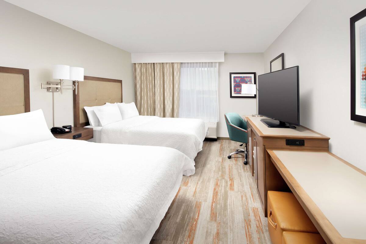 A hotel room with two beds, a desk, a television, and a window with curtains.
