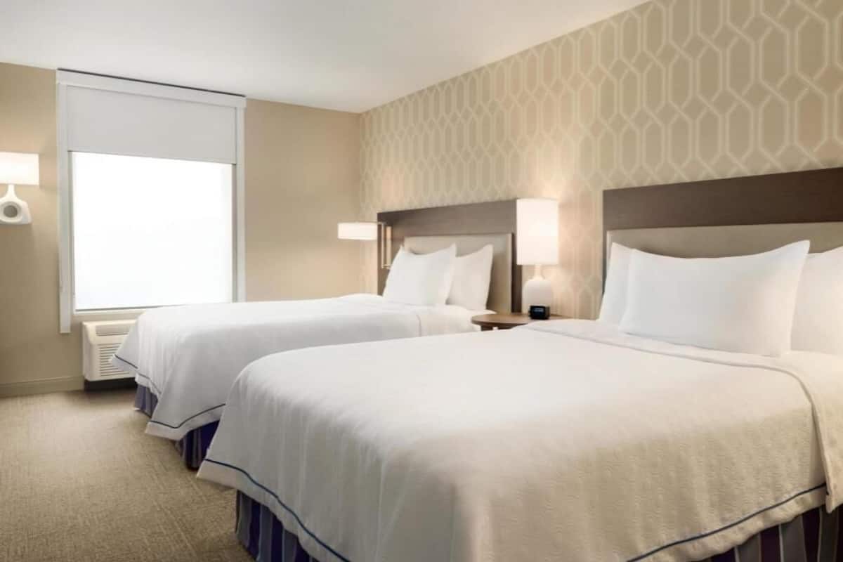 A hotel room with two beds, a window with blinds, a wall with a geometric pattern, and bedside lamps.