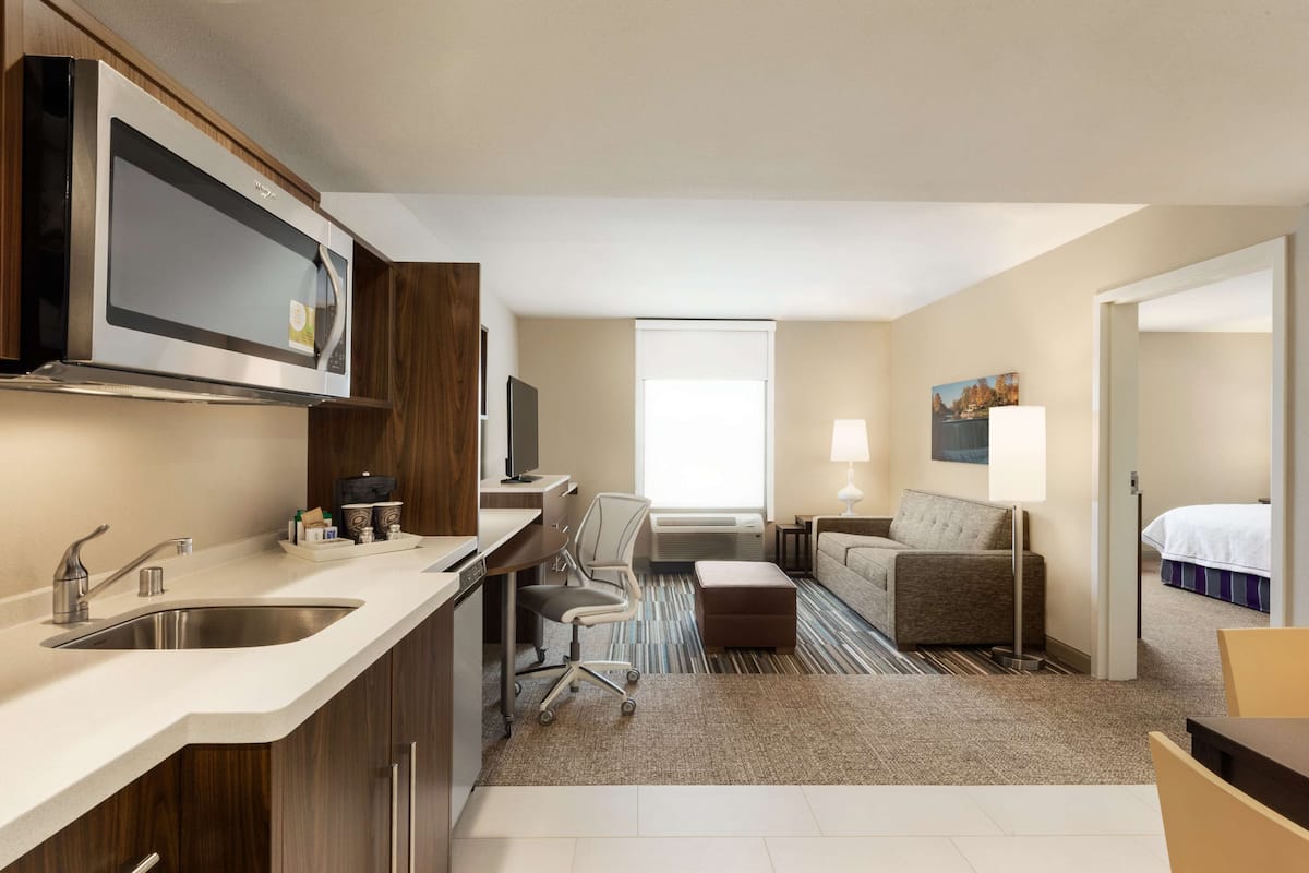 A compact hotel room with a kitchenette, a sofa, a bed, and a window.