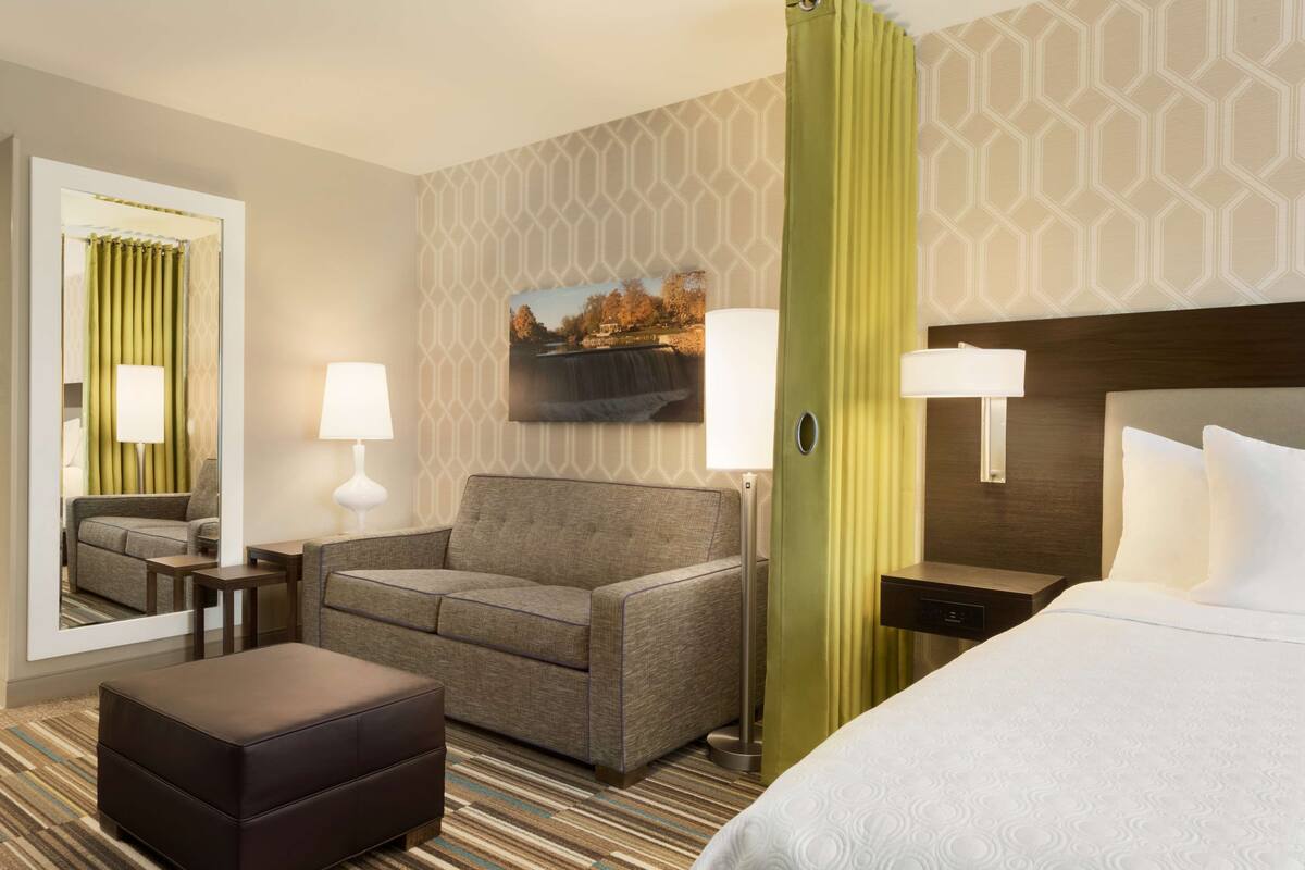 A hotel room with a sofa, ottoman, a bed, a nightstand, a lamp, a mirror, and a picture on the wall.