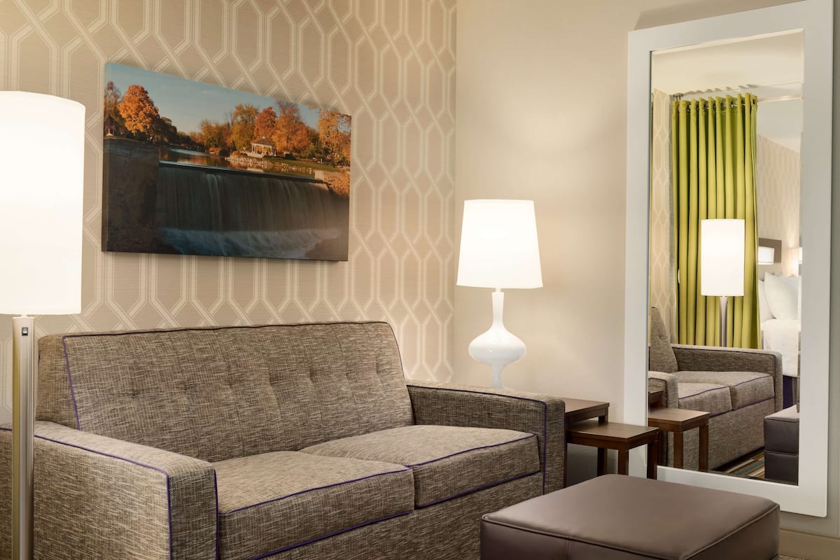 A hotel room with a sofa, a small table, a lamp, a painting of a waterfall, and a mirror reflecting the room.