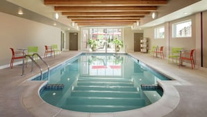 Una piscina interior con vistas despejadas al exterior, rodeada de sillas verdes y rojas, y vigas de madera en el techo.