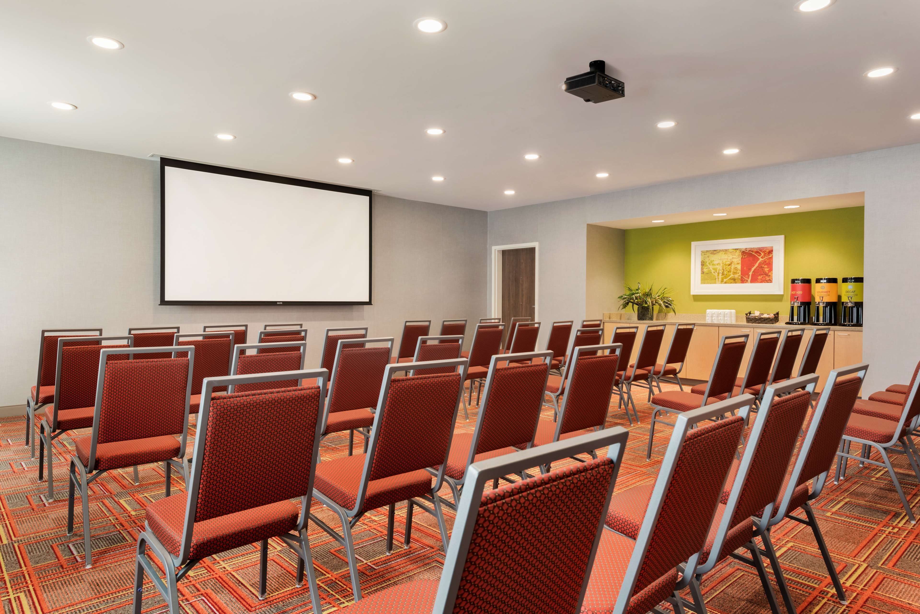 Una sala de conferències amb files de cadires vermelles, una pantalla gran i una taulella de recepció amb una planta.