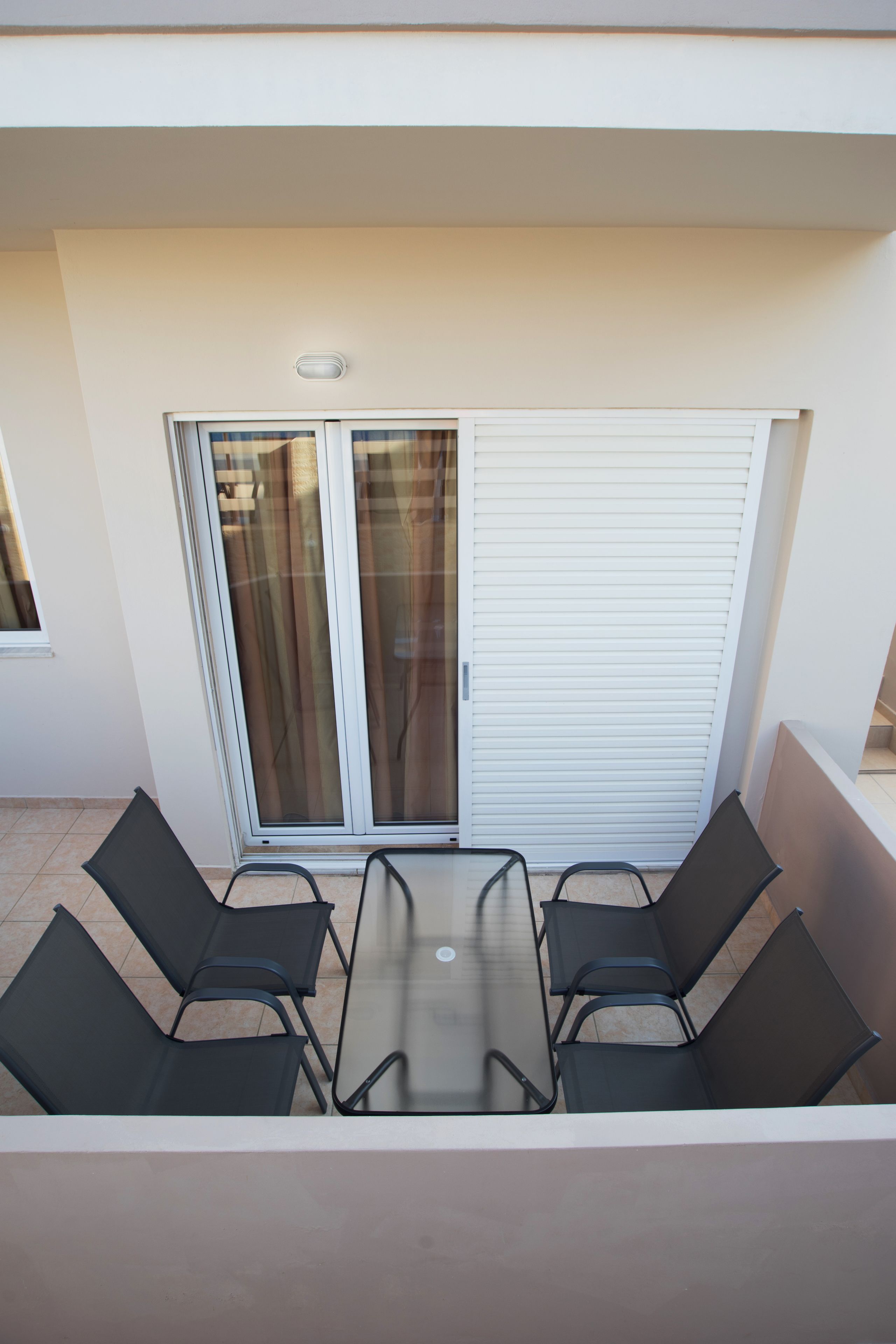 A balcony with a glass table and four chairs, a sliding glass door, and a white exterior wall.