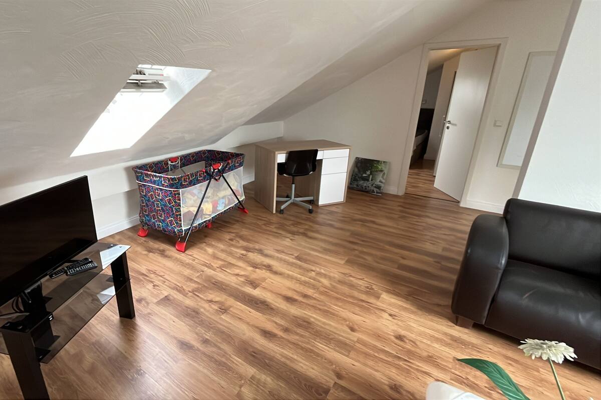 Een modern loft appartement met een schuine kap, houten vloer, een zwarte leren bank, een televisie en een kleurrijke kinderwagen.