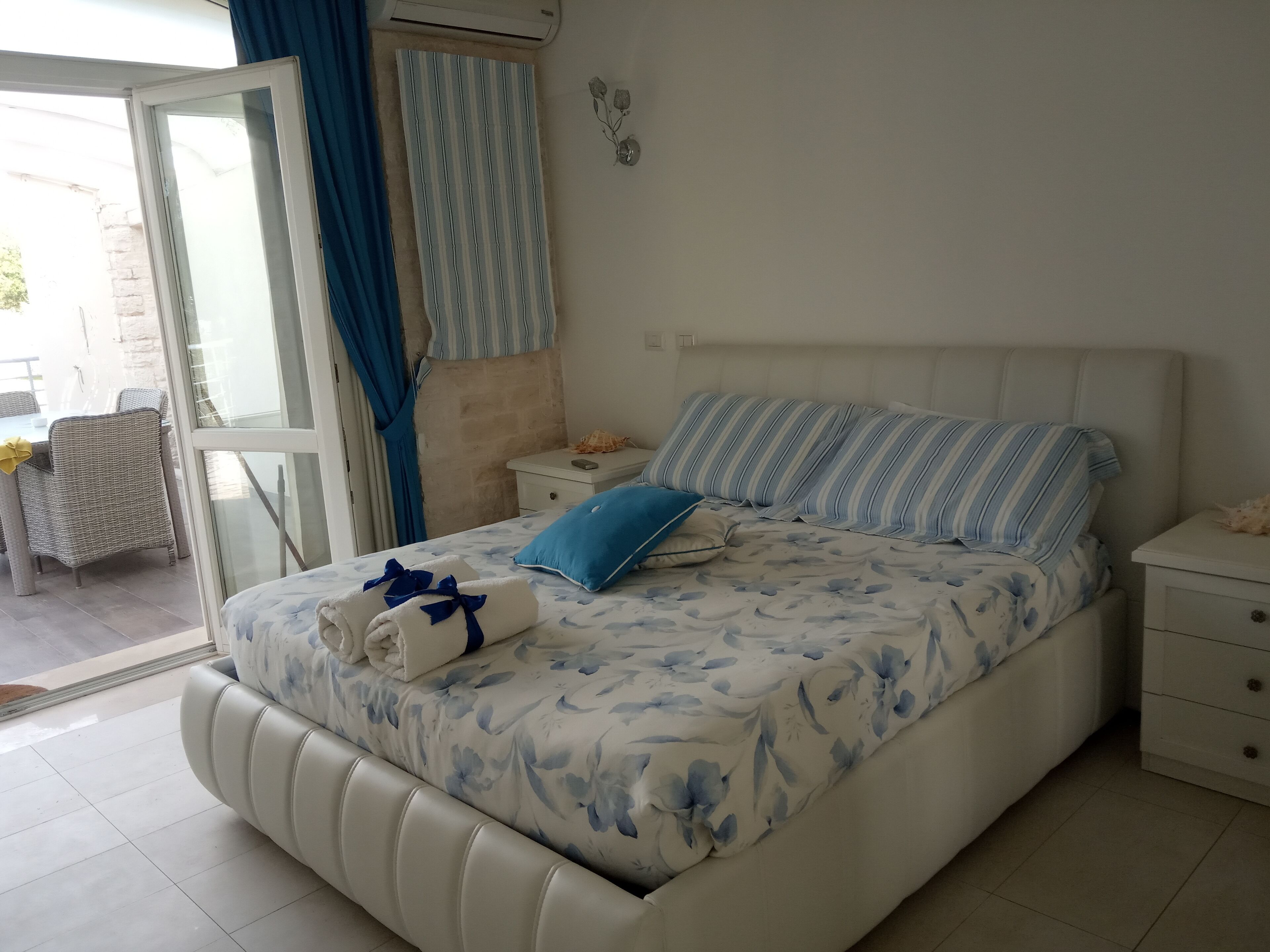 A bedroom with a bed, a nightstand, a window with curtains, and a view of the outside.