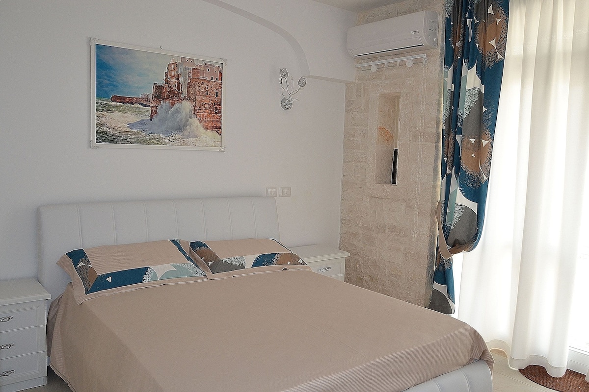 Une chambre à coucher comprenant un lit, une table de chevet, un climatiseur fixé au mur et un tableau représentant un paysage côtier.