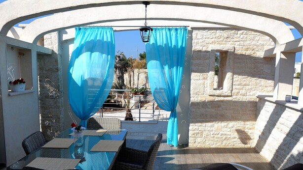 An outdoor dining area with wicker chairs and a glass table, blue curtains, and a stone wall backdrop.