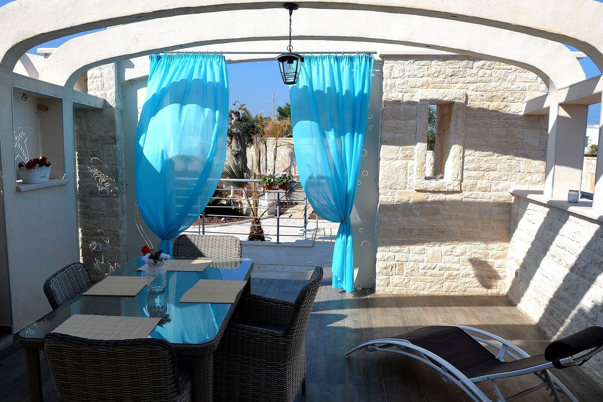 Un espace de restauration en plein air avec des chaises en osier et une table en verre, des rideaux bleus et un mur en pierre en arrière-plan.