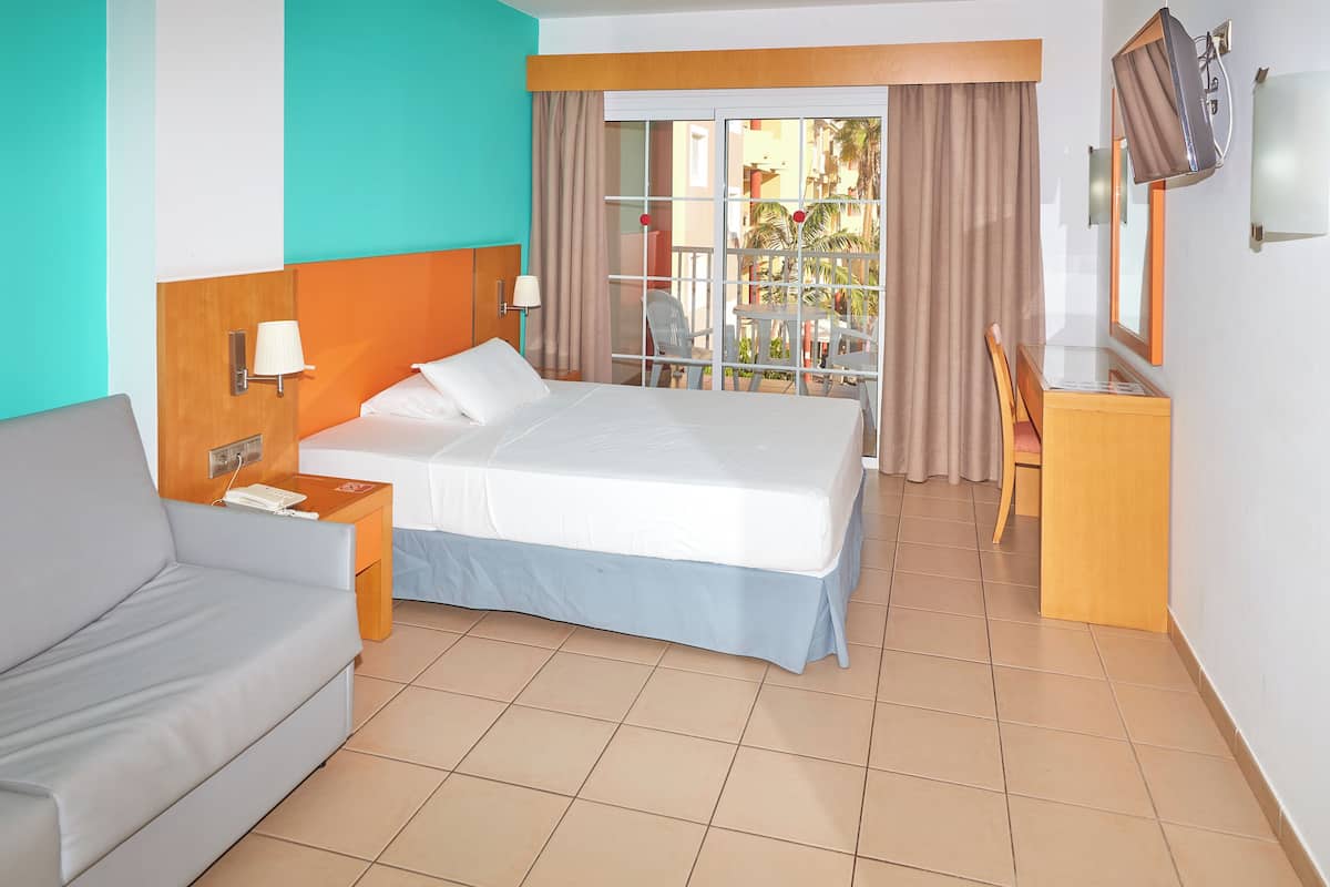 A hotel room with a bed, a sofa, a desk, a TV, and a balcony with a view of palm trees.