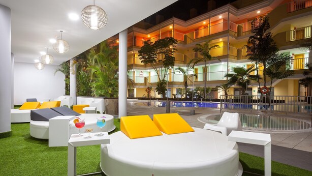 Un área de estar exterior moderna con una disposición circular de asientos, una piscina y un edificio con balcones al fondo.