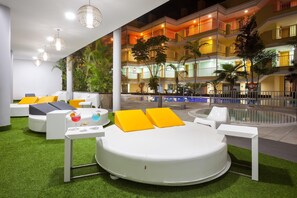 A modern outdoor lounge area with a circular seating arrangement, a pool, and a building with balconies in the background. - Hotel Chatur Costa Caleta (Antigua)
