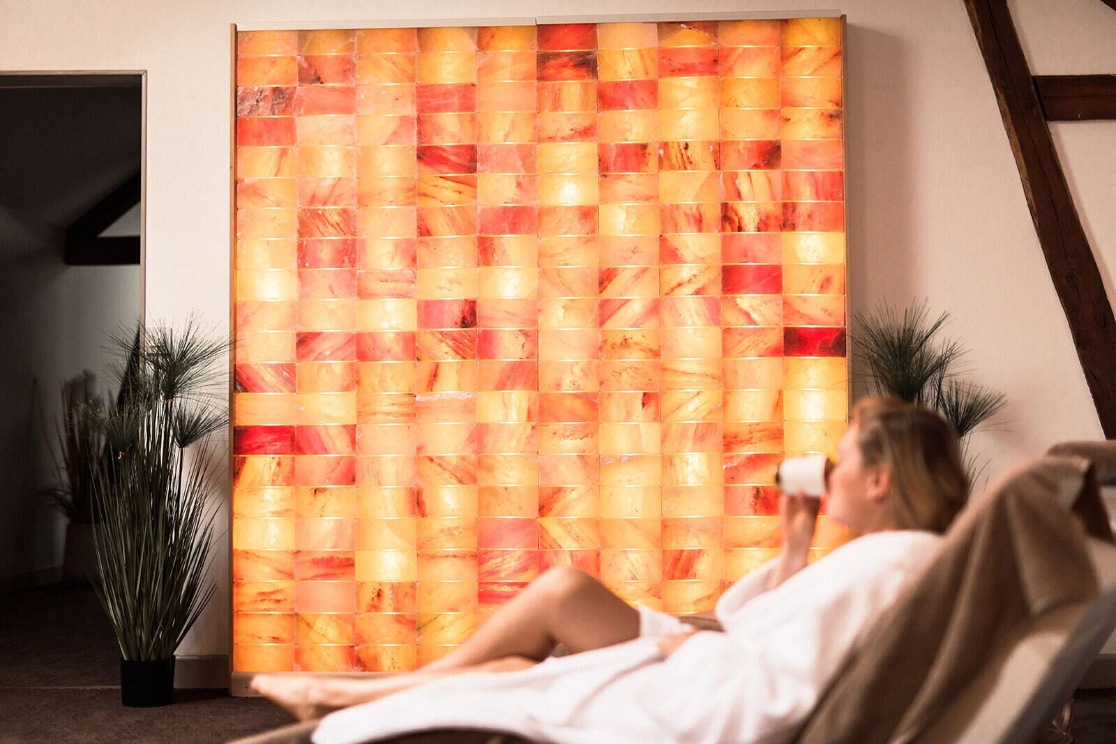 A room with a large, textured wall panel, a potted plant, and a person relaxing in a chair.