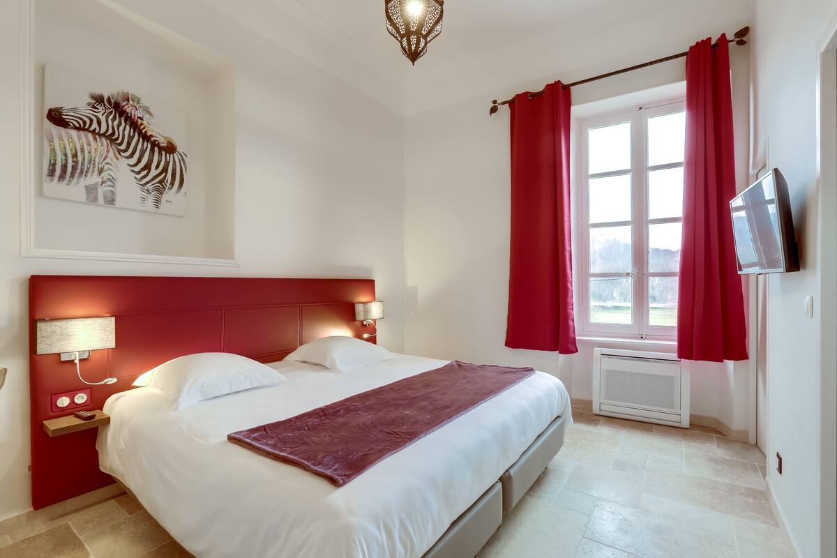 Habitación de hotel con cama, televisor montado, ventana con cortinas rojas, cuadro de una cebra en la pared y lámpara de noche.