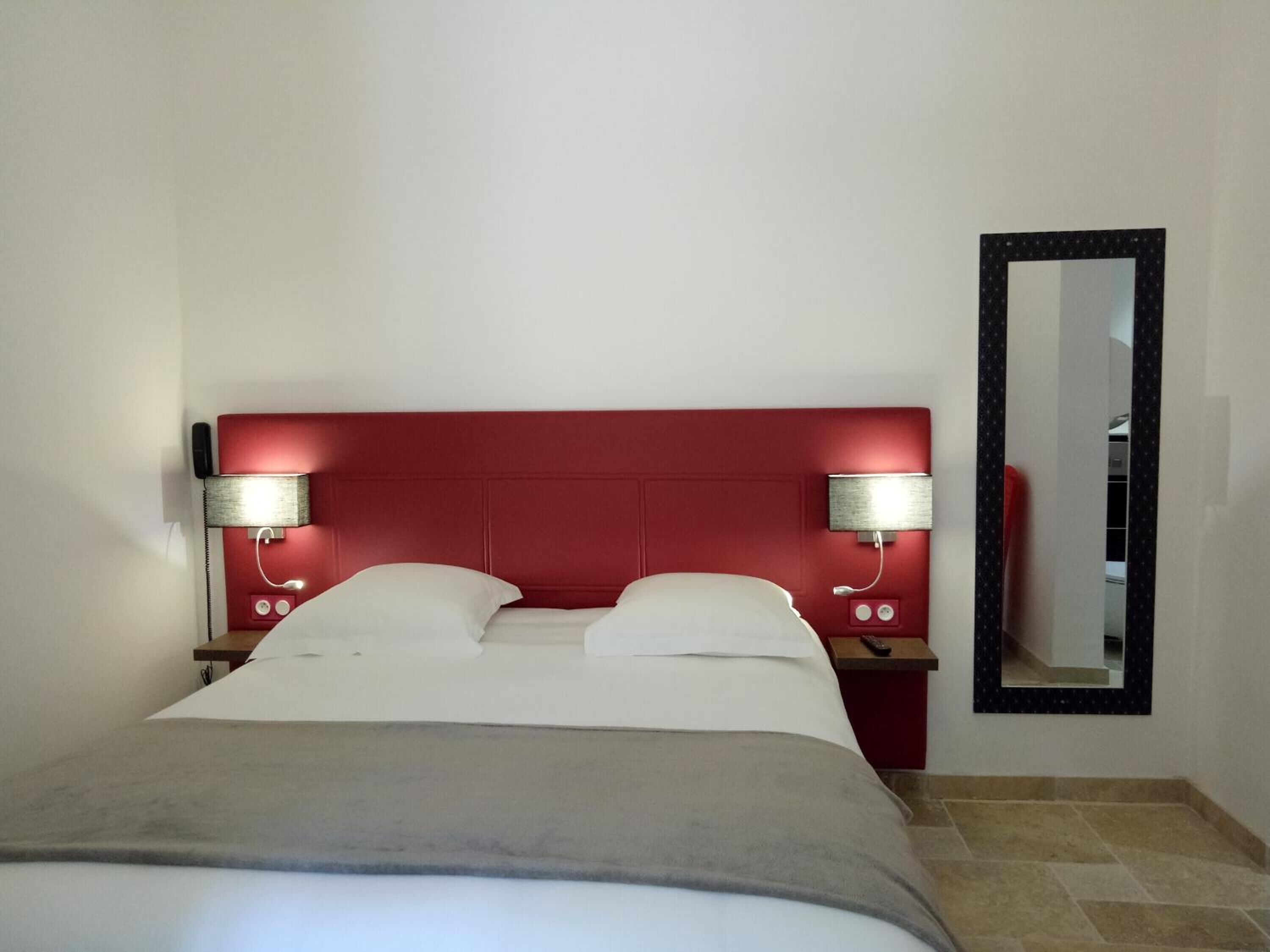 Une chambre d’hôtel avec une tête de lit rouge, deux lampes de chevet, un miroir et un lit recouvert de draps blancs.