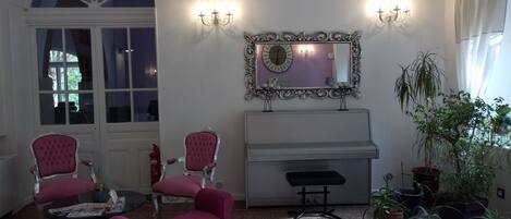 A room with a piano, two pink chairs, a bench, a mirror, and several potted plants.