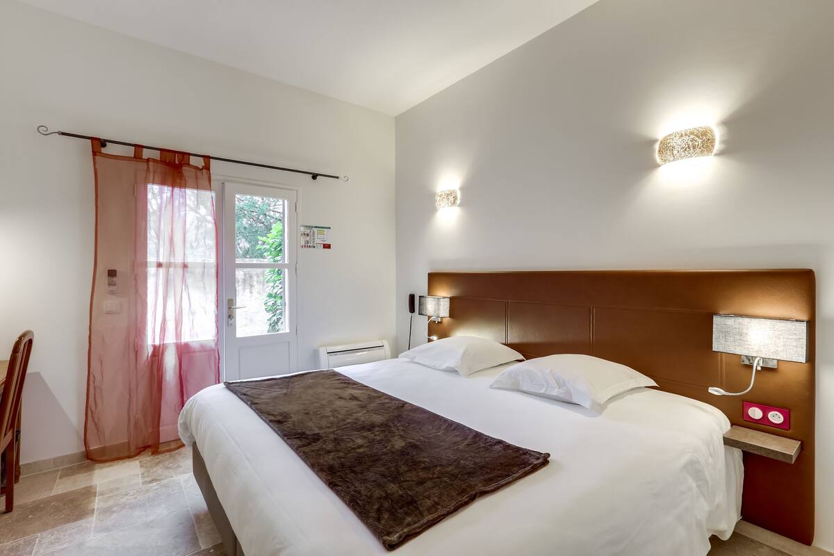 Habitación de hotel con una cama grande, cabecera de madera, dos lámparas de noche, una mesita de noche y una ventana con cortinas traslúcidas.