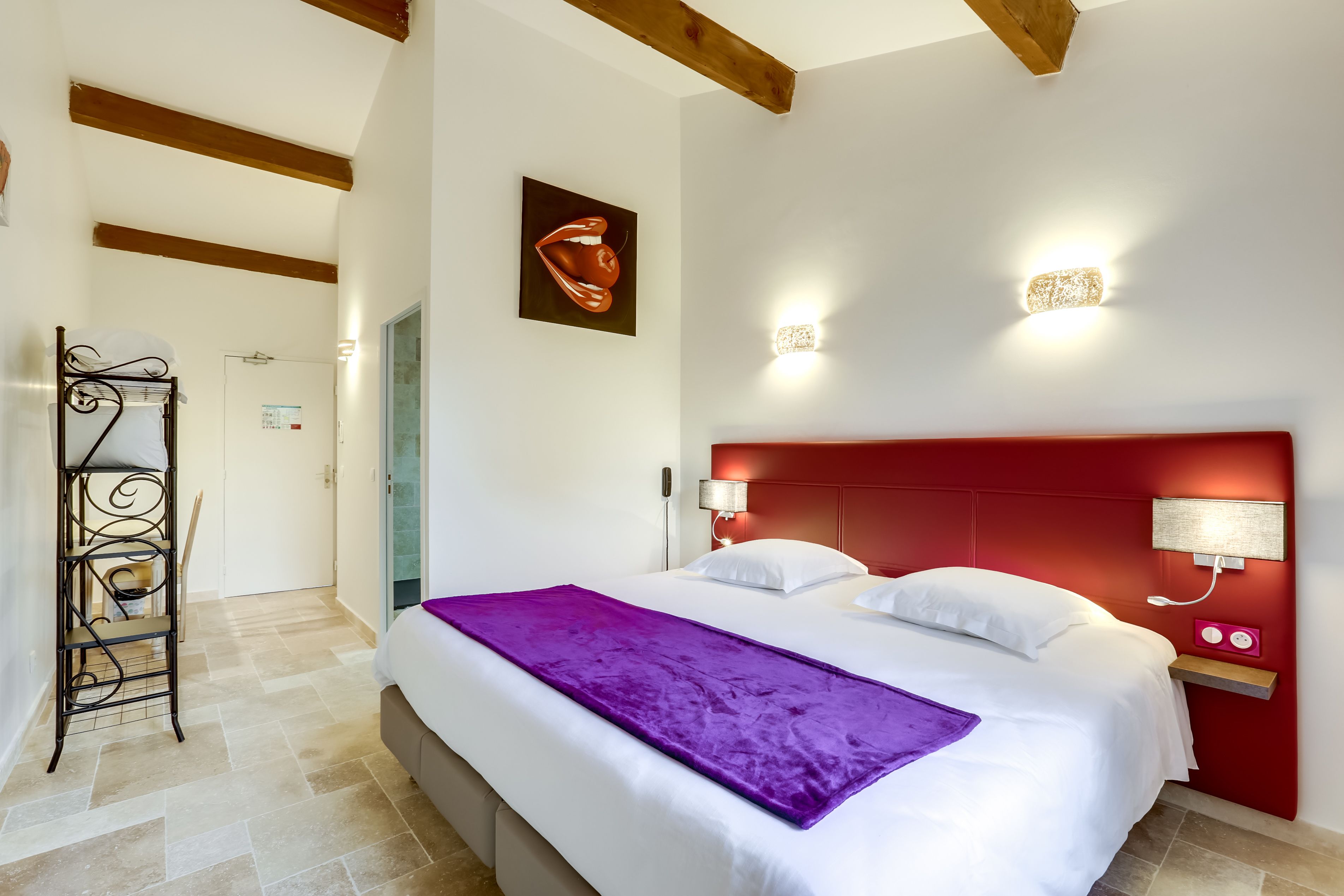 Quarto de hotel com cama, cabeceira vermelha, manta roxa e luminária fixa na parede.