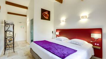 Quarto de hotel com cama, cabeceira vermelha, manta roxa e luminária fixa na parede.