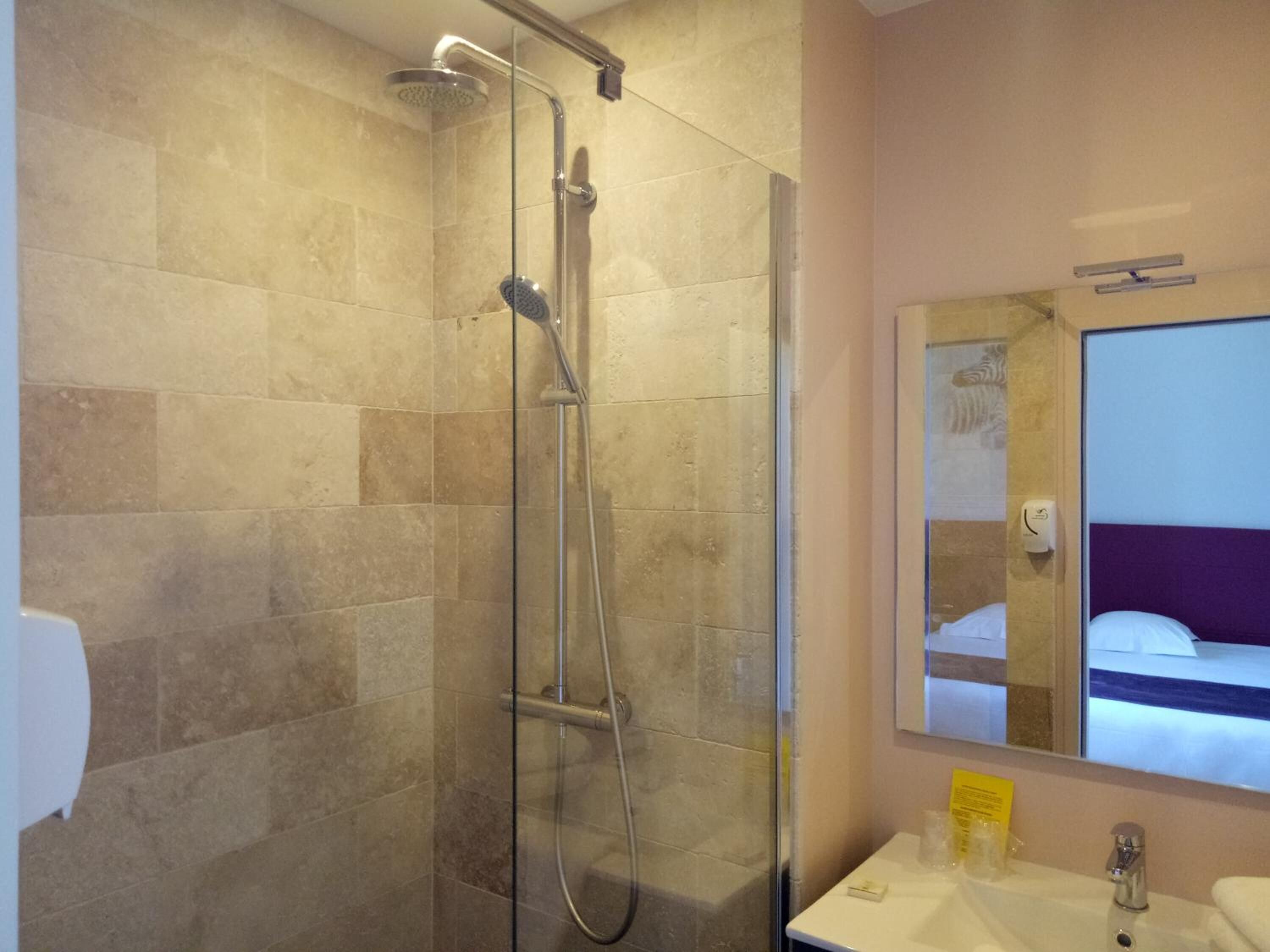 A bathroom with a glass-enclosed shower, a white sink, and a mirror reflecting a bed.
