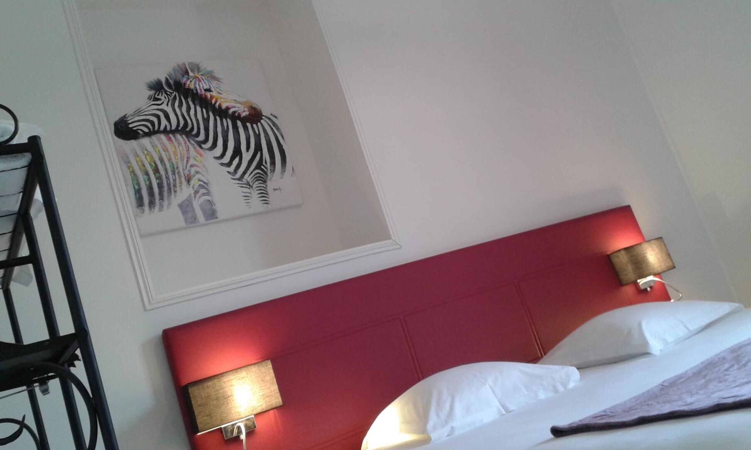 Una habitación de hotel con cabecera roja, dos lámparas de noche, un cuadro de cebra y una cama con ropa blanca.
