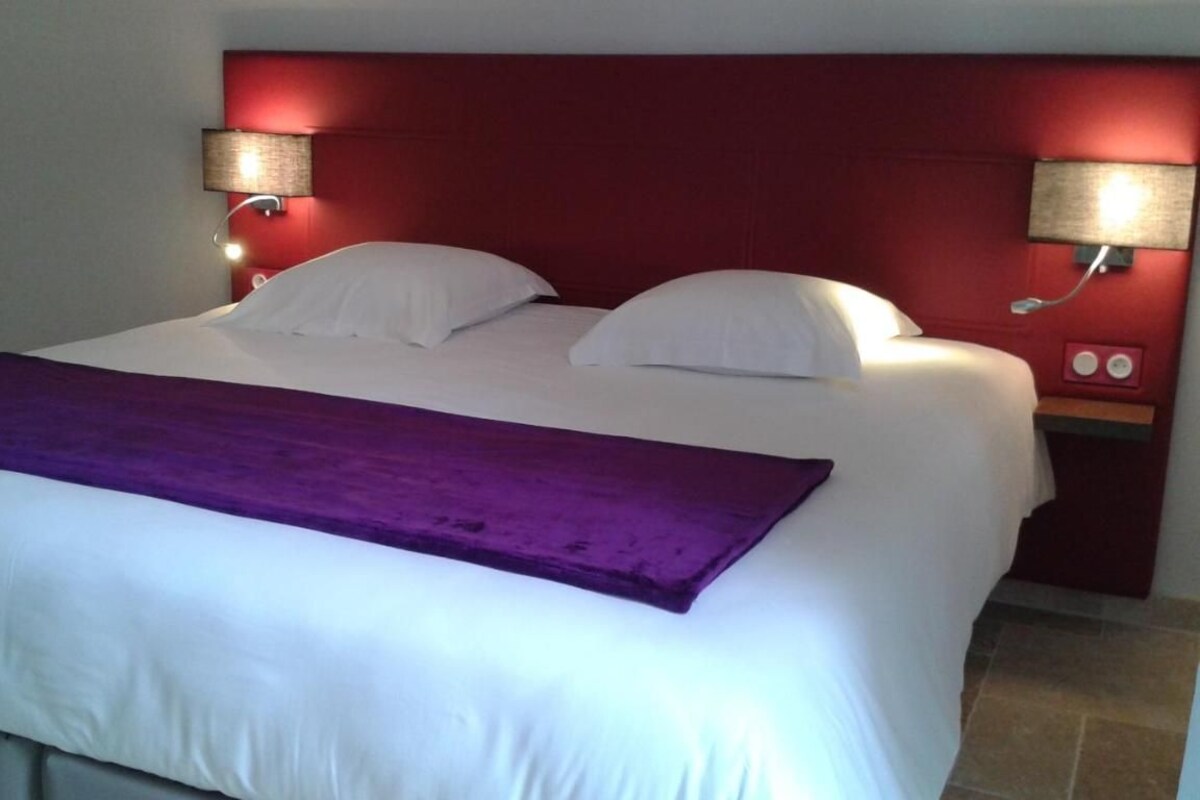Une chambre d’hôtel avec un grand lit, deux lampes de chevet, une tête de lit rouge et un plaid violet.