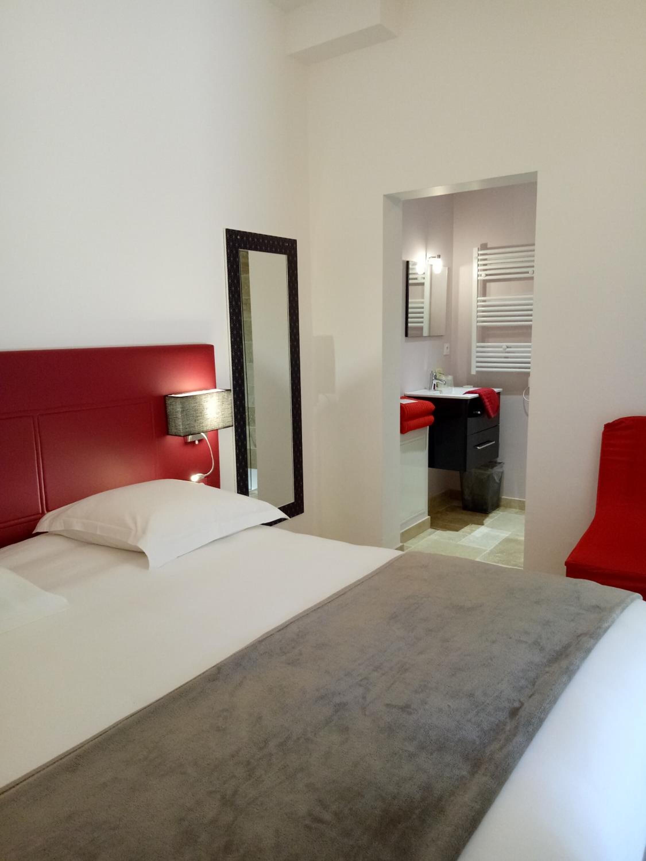 Una habitación de hotel con cama, cabecero rojo, lámpara de noche, espejo grande y silla roja.