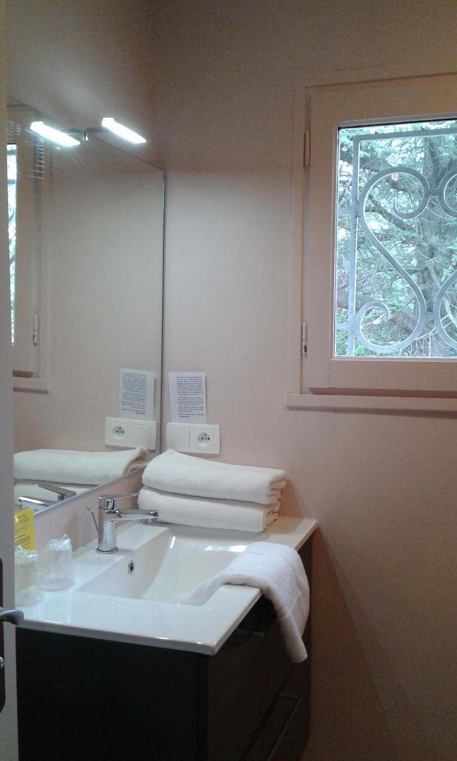 Une salle de bain avec un lavabo blanc, un miroir et une fenêtre ornée d’un cadre en métal décoratif. Deux panneaux sont accrochés au mur, l’un au-dessus du lavabo et l’autre en dessous.