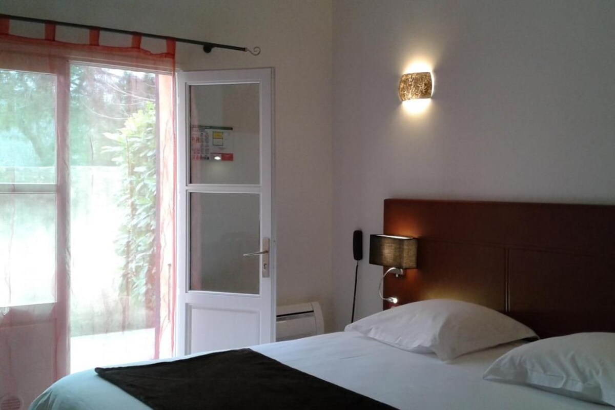 A hotel room with a bed, a nightstand with a lamp, a door with a view to the outside, and a wall-mounted light fixture.