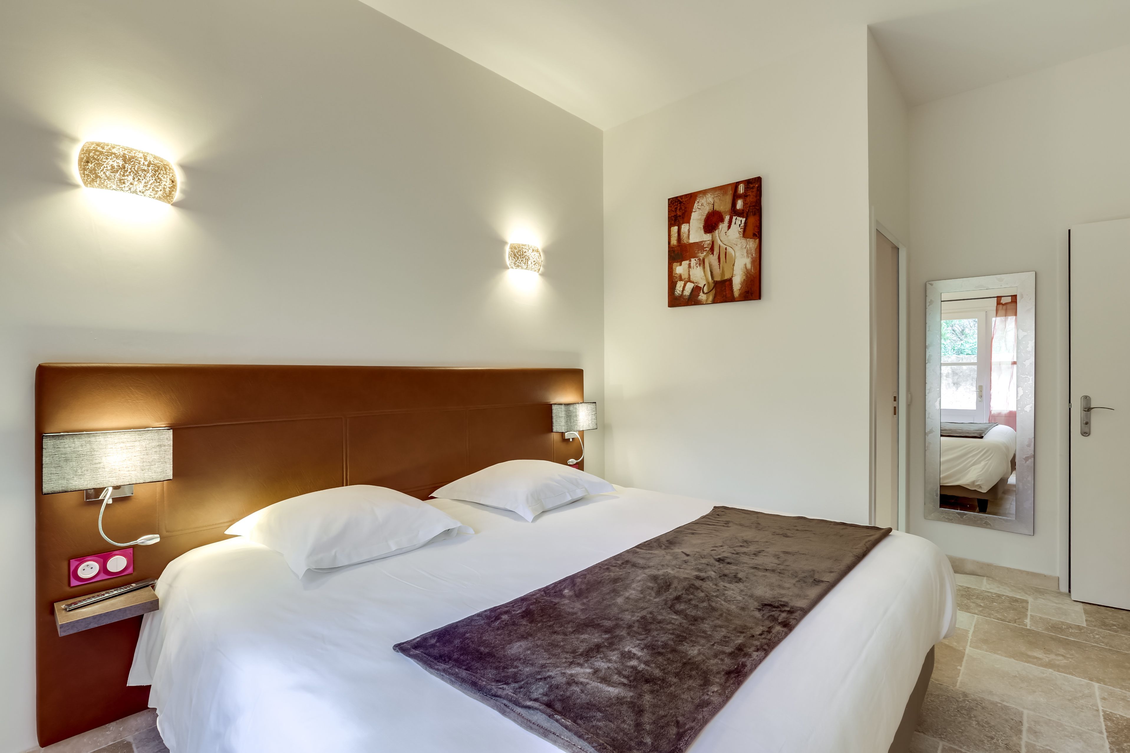 Habitación de hotel con una cama grande, cabecera, lámparas de noche, mesitas de noche, un espejo y un cuadro en la pared.