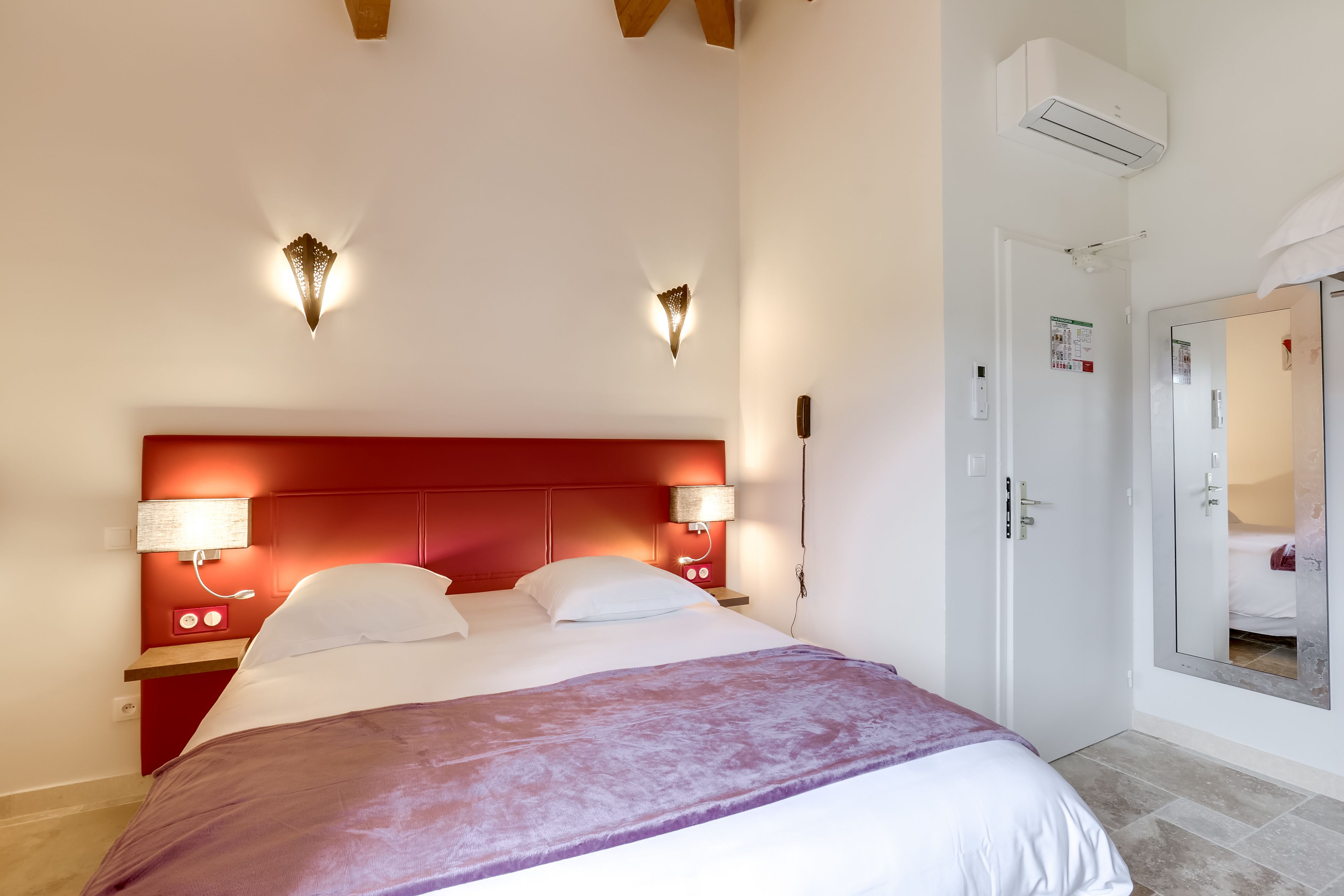 Habitación de hotel con cabecera roja, una cama grande con ropa de cama blanca, dos lámparas de noche y un equipo de aire acondicionado en la pared.