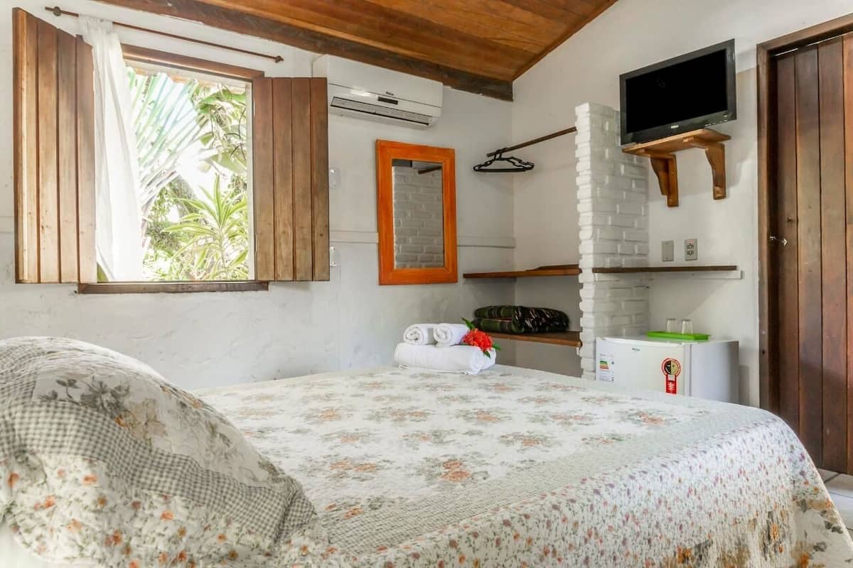 A bedroom with a bed, a TV mounted on a brick wall, an air conditioning unit, and a window with wooden shutters.