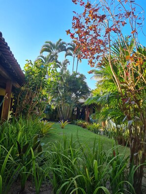 A lush garden with various tropical plants and palm trees, a wooden structure, and a clear blue sky. - Pousada Varanda do Sol (Porto Seguro)