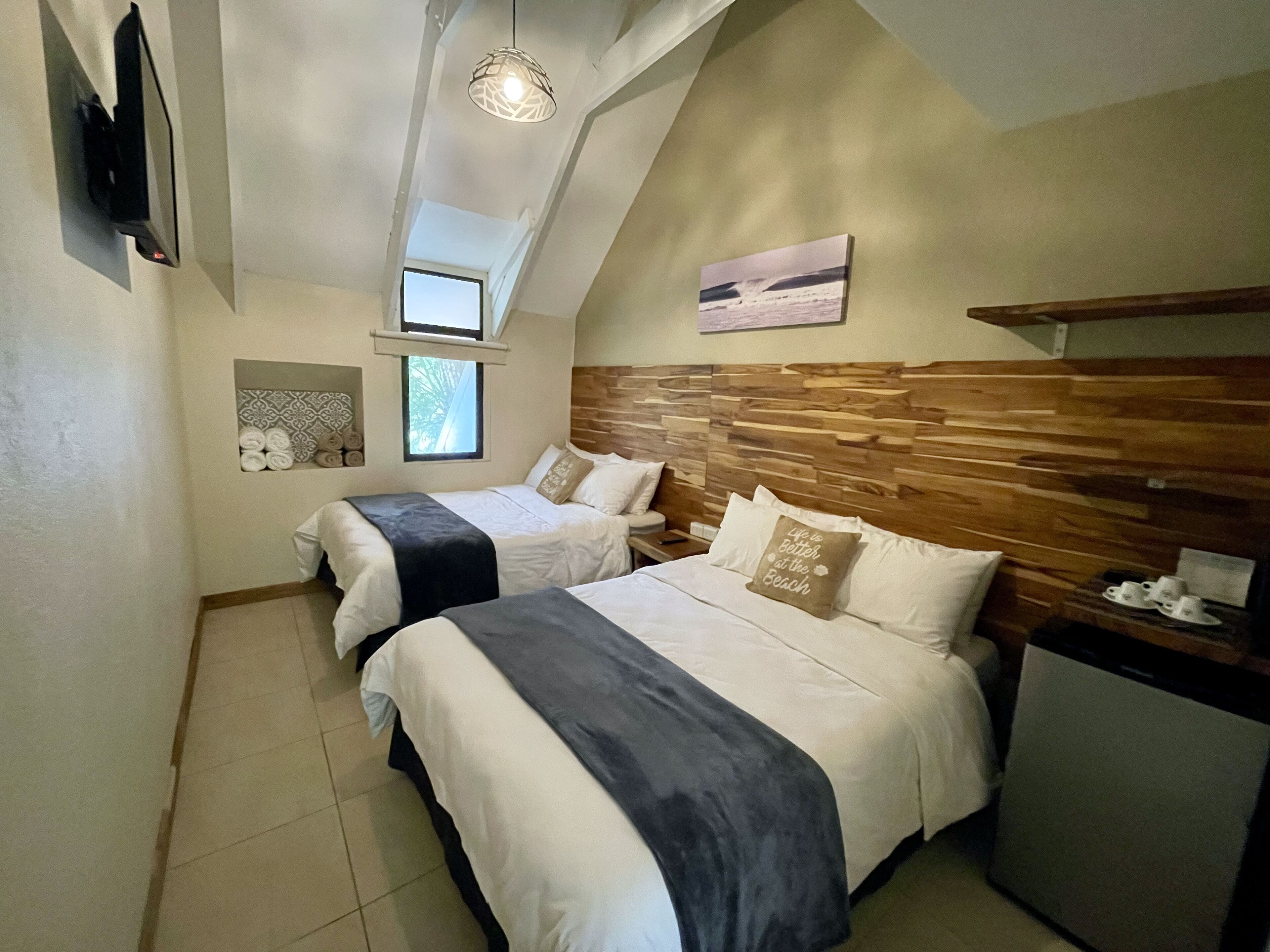 A bedroom with two beds, a wooden headboard, a small window, a TV, and a shelf with cups.