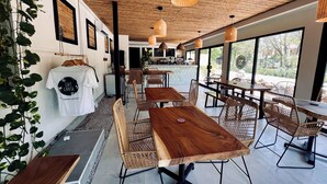 Ein Café mit Holztischen, Korbstühlen, einer Bar und großen Fenstern mit Blick ins Freie.