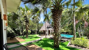 Eine Holzterrasse mit Stuhl, einem Gebäude mit Strohdach und einem Swimmingpool, der von  üppiger Vegetation umgeben ist.