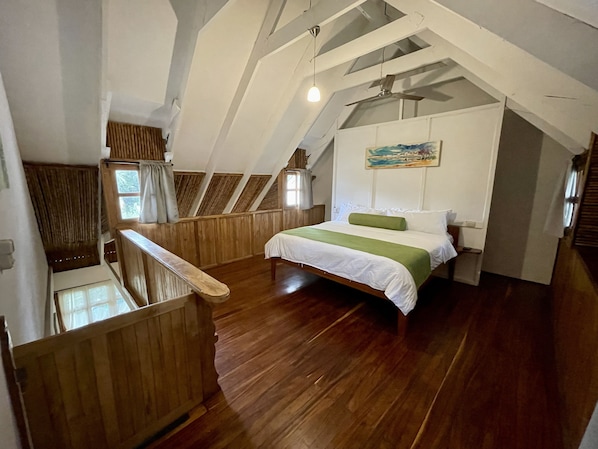 A bedroom with a wooden floor, a bed with a green and white bedspread, a painting on the wall, and a window with curtains. - Hotel El Manglar & Cantarana (Cabo Velas)