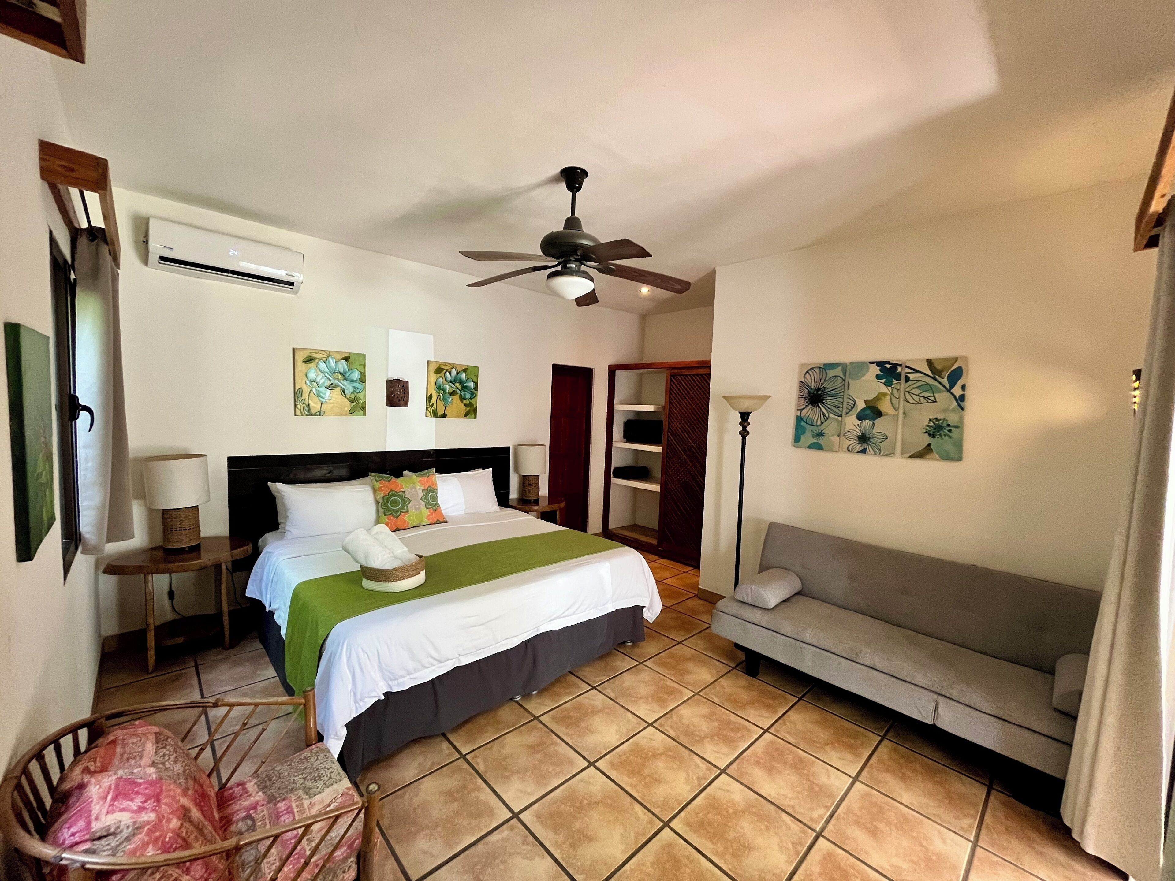 A hotel room with a bed, a sofa, a ceiling fan, artwork on the walls, and a small table with a lamp.