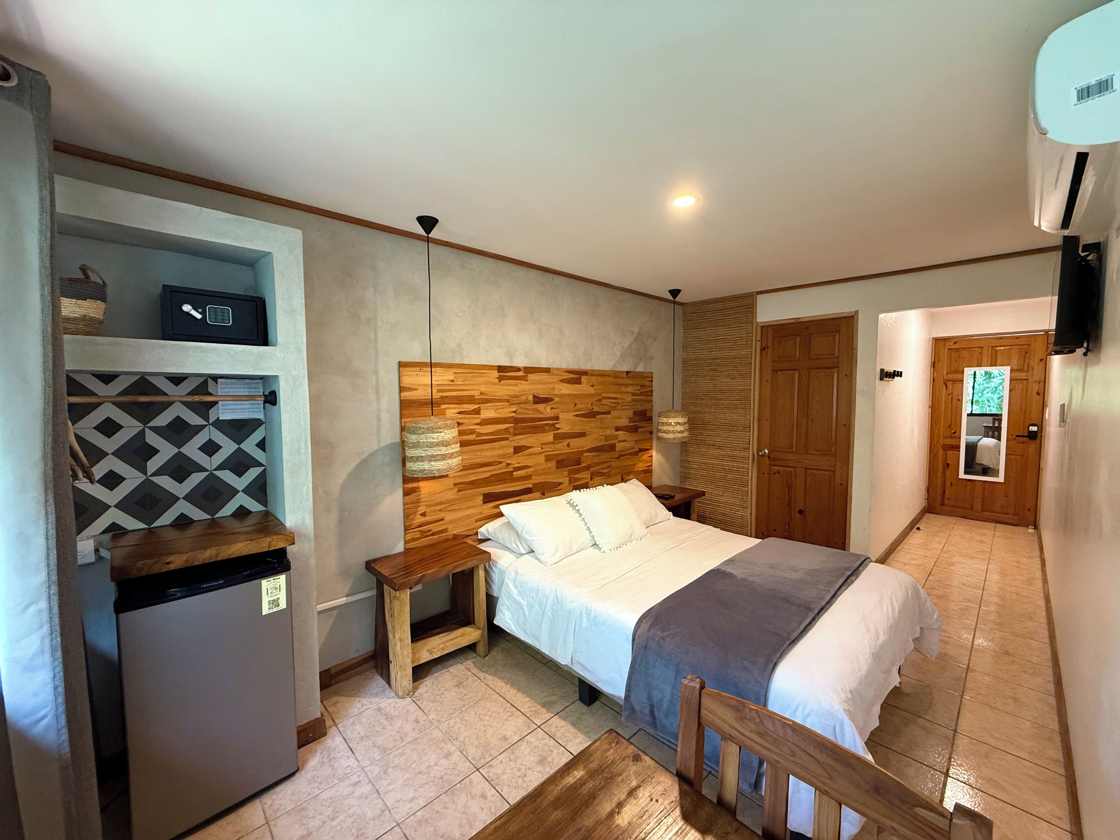 A bedroom with a bed, bedside tables, a wooden headboard, a wall-mounted air conditioner, and a door with a mirror.