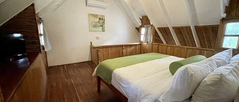 A bedroom with a sloped ceiling, wooden flooring, a bed with white bedding, a green throw pillow, and a painting on the wall.