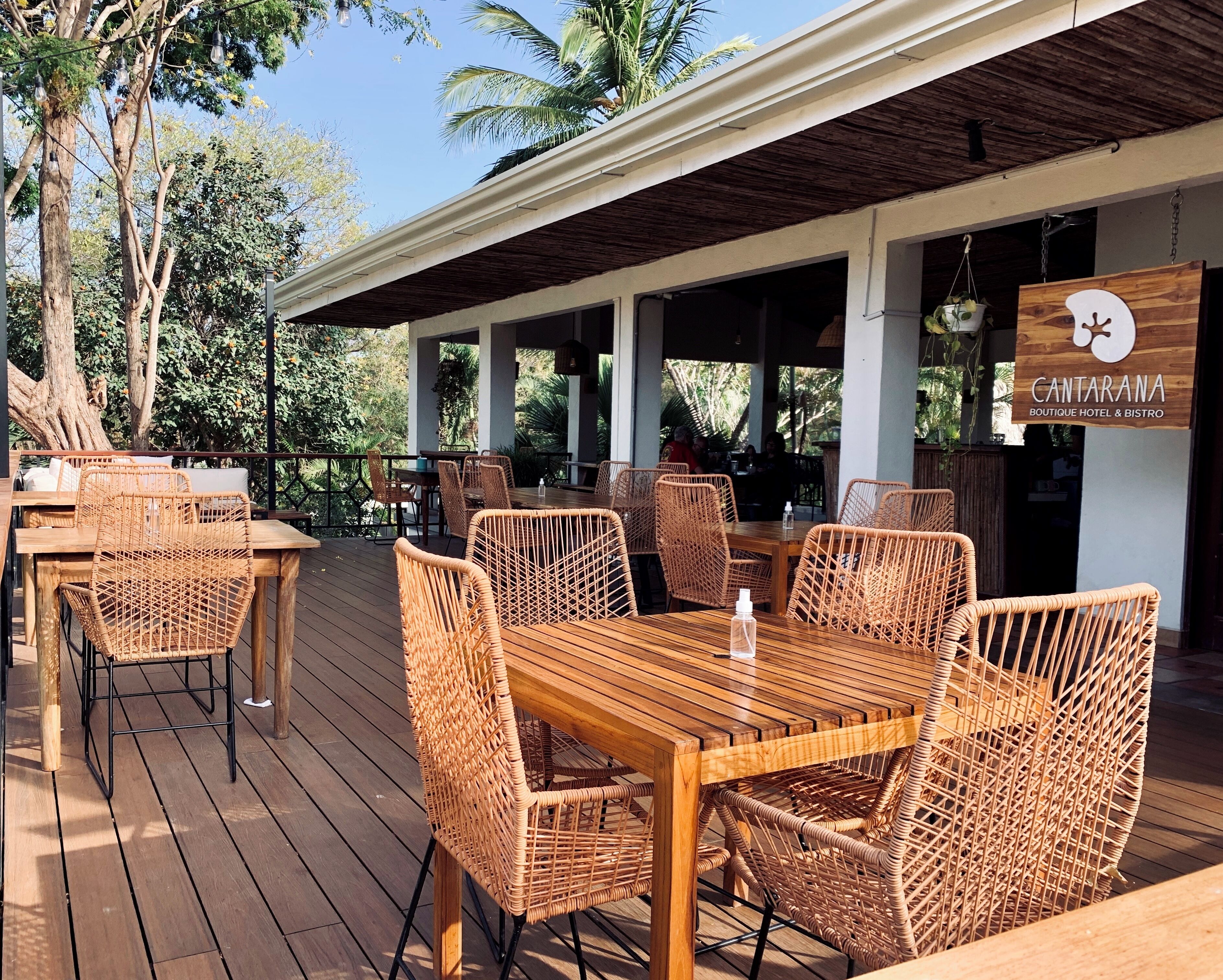An outdoor dining area with wicker furniture and a wooden table, surrounded by lush greenery and a sign for Cantarana Boutique Hotel & Bistro.