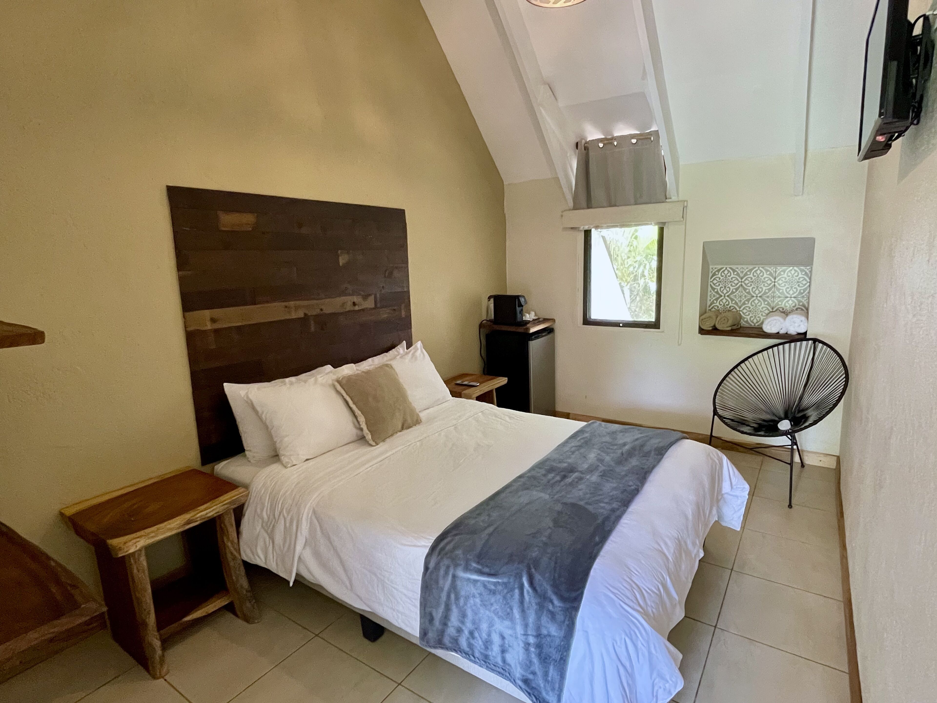Une chambre à coucher avec une tête de lit en bois, un lit recouvert de draps blancs, une table de chevet, une chaise, un petit bureau avec une lampe et une fenêtre donnant sur un paysage verdoyant.