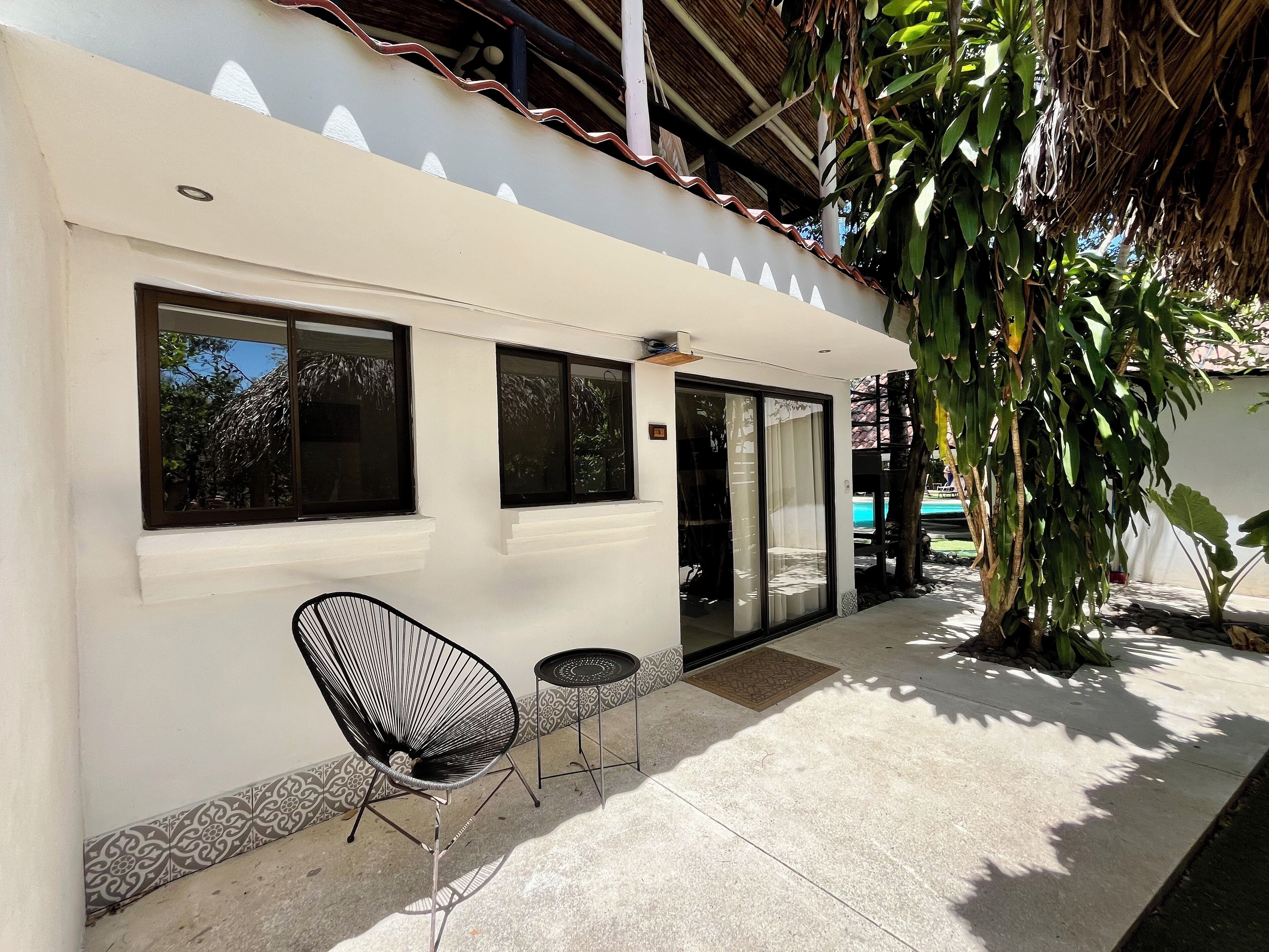 A modern building with large windows, a covered patio area with a chair and table, and a large tree providing shade.