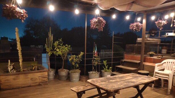 Een terras buiten met een houten tafel en banken, omringd door potplanten en bloemenmanden.