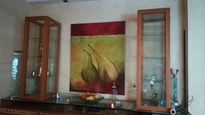 Een eethoek met een tafel met glazen blad, twee vitrinekasten met houten frame, en een schilderij van peren aan de muur.