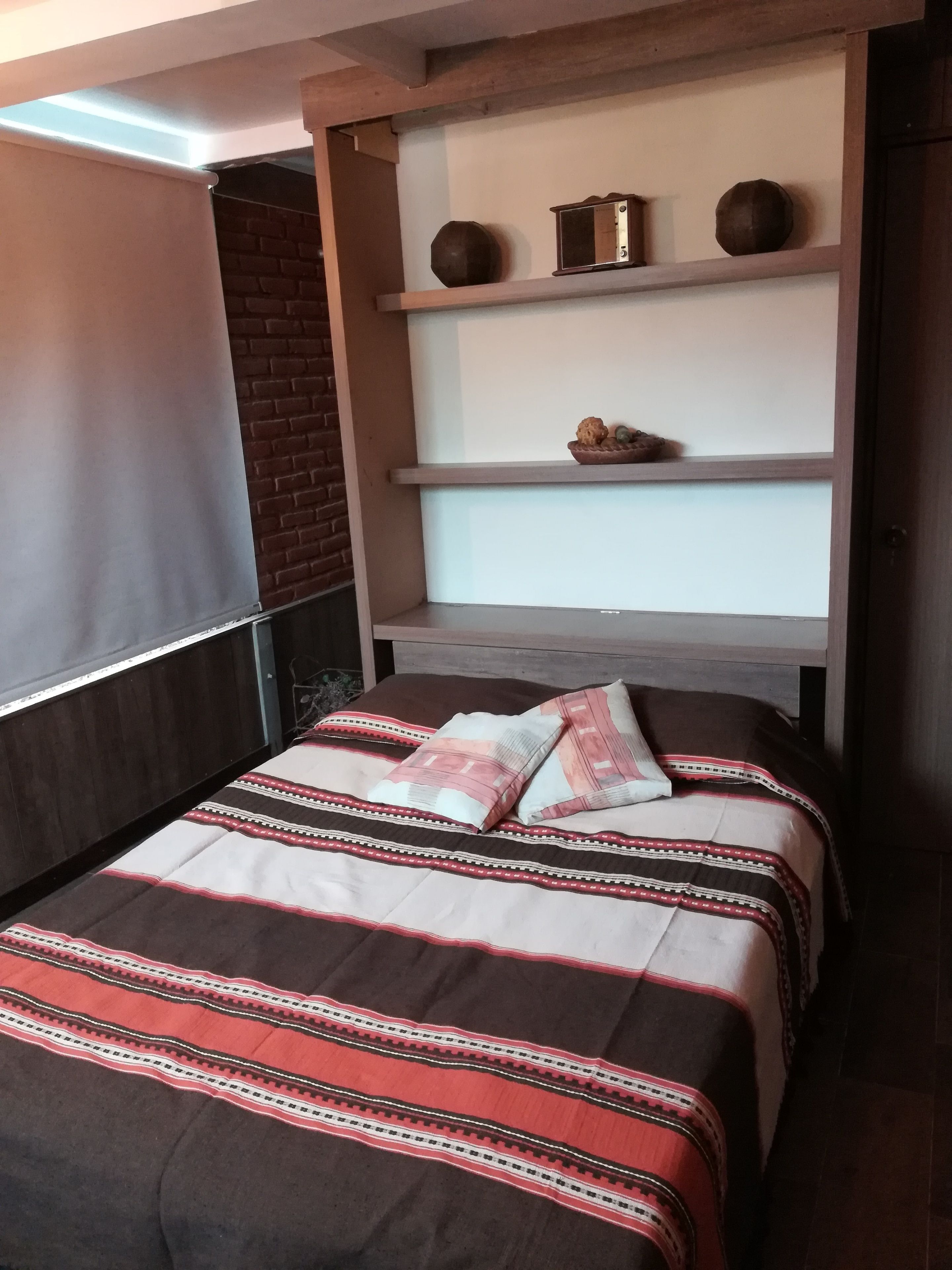 Une chambre à coucher avec un lit, un mur en briques, une étagère ornée d’objets décoratifs et une fenêtre.
