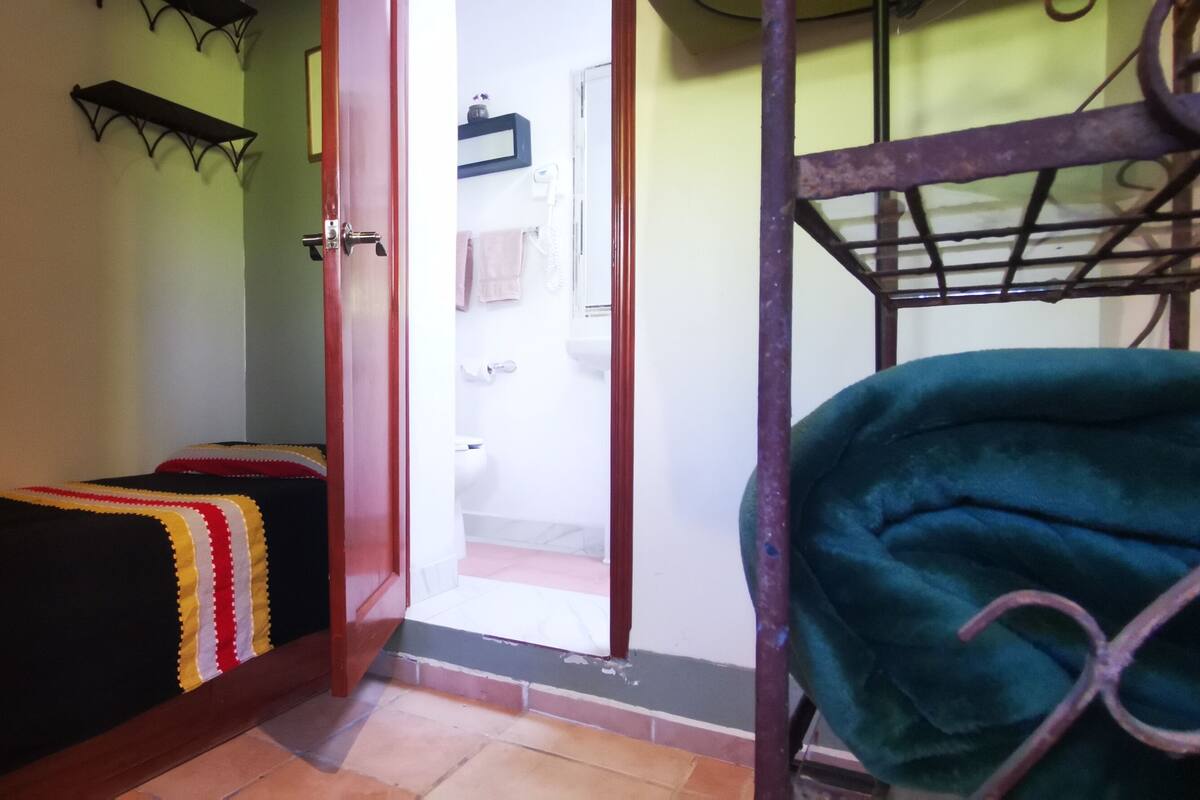 Une pièce comprenant un lit, un banc, une étagère en métal et une porte donnant sur une salle de bain.