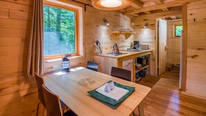 A wooden cabin interior with a dining table, chairs, a kitchen area, and a large window with curtains. - Huttopia White Mountains (Conway)